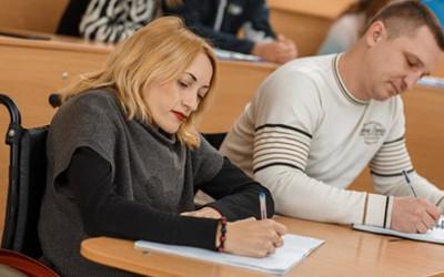 A lady wearing dark clothes, sitting in a wheelchair at a desk next to a man wearing a white top, both of them writing on notepads