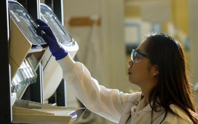 Scientist with long, dark hair, wearing safety glasses, operating laboratory equipment