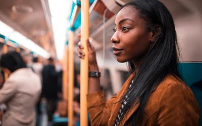 A young, black girl, with long dark hair and wearing a brown jacket, standing on a train and holding on to a rail