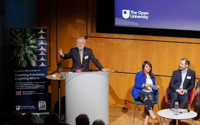 Professor Nick Braithwaite on stage in front of a lecturn, with a man and a woman sitting to his left, and with a big screen in the background