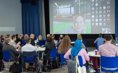 Hybrid meeting, with a group of people in a room and screen showing people attending by video
