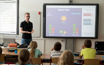 A male teacher standing in front of school children sitting at desks, with a monitor on the wall 