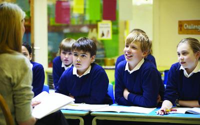 A teacher reading to a group of school children who are wearing white collared shirts and blue jumpers