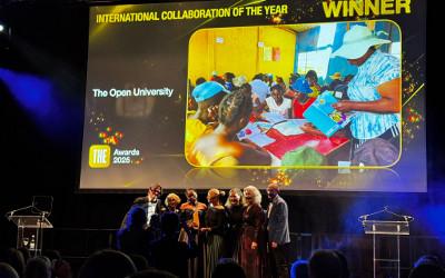 A group of people on a stage collecting an award, with a a screen behind them showing a picture in a room sitting at tables with books, and the words The Open University, International Collaboration of the Year, Winner, THE Awards 2025