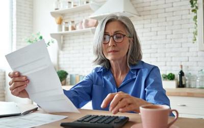 Woman wearing glasses, sitting at a table with a piece of paper in her hand and a calculator and coffee cup on the table