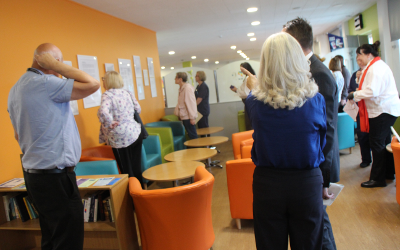 A group of people in a room with small round tables, green, blue and orange tub chairs, looking at notices on an orange wall
