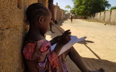 A young African girl sitting in a dusty street with a book and pen
